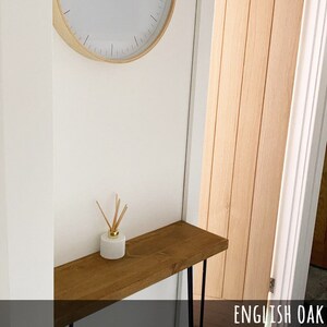 May include: A wooden console table with black hairpin legs, a white diffuser with reeds, and a wall clock. The table is made of English oak. The clock has a light wood frame and a white face.