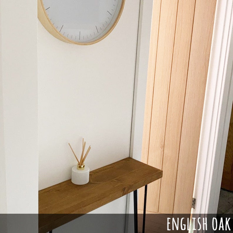 May include: A wooden console table with black hairpin legs, a white diffuser with reeds, and a wall clock. The table is made of English oak. The clock has a light wood frame and a white face.