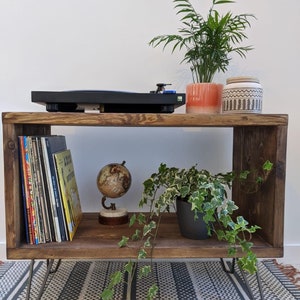 May include: A rustic wooden shelf with hairpin legs, featuring a record player, a globe, and two potted plants. The shelf has two open shelves, one with a collection of vinyl records and the other with a potted plant. The shelf is sitting on a striped rug.