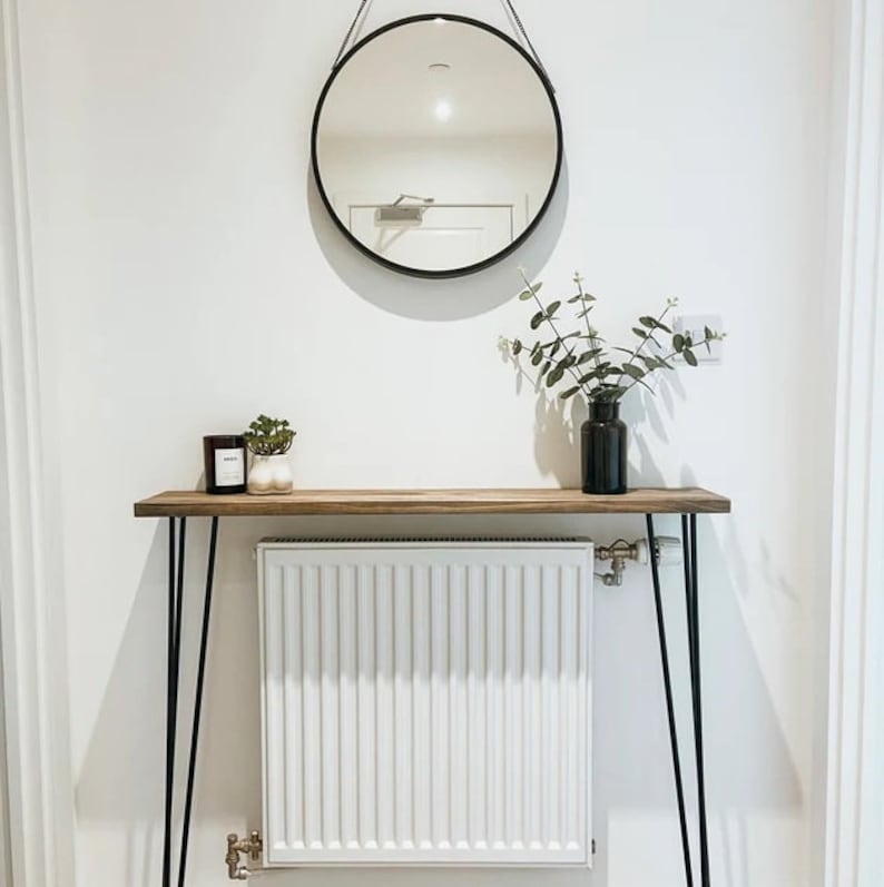 May include: A round black mirror hangs above a wooden console table with black hairpin legs. The table holds a black vase with greenery, a candle, and a small plant. A white radiator is below the table.