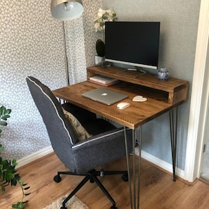 May include: A wooden desk with a metal hairpin leg design. The desk has a shelf above the desktop and a laptop computer, a mouse, and a white heart on the desktop. The desk is in a room with a patterned wallpaper and a floor lamp.