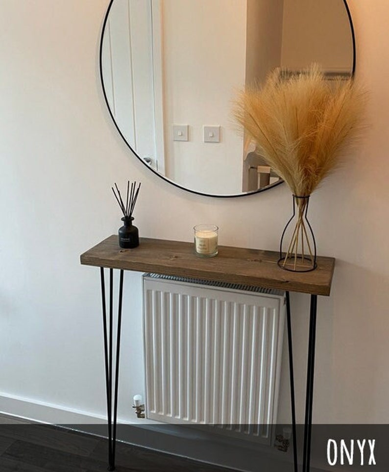 May include: A rustic wooden console table with black hairpin legs. The table is adorned with a black diffuser, a lit candle in a glass jar, and a decorative vase holding dried pampas grass. A round black-framed mirror hangs on the wall.