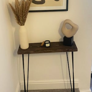May include: A dark brown wooden console table with black hairpin legs. The table is decorated with a beige vase holding dried plants, a small candle, and a beige abstract sculpture on a black base. A framed abstract artwork hangs above.