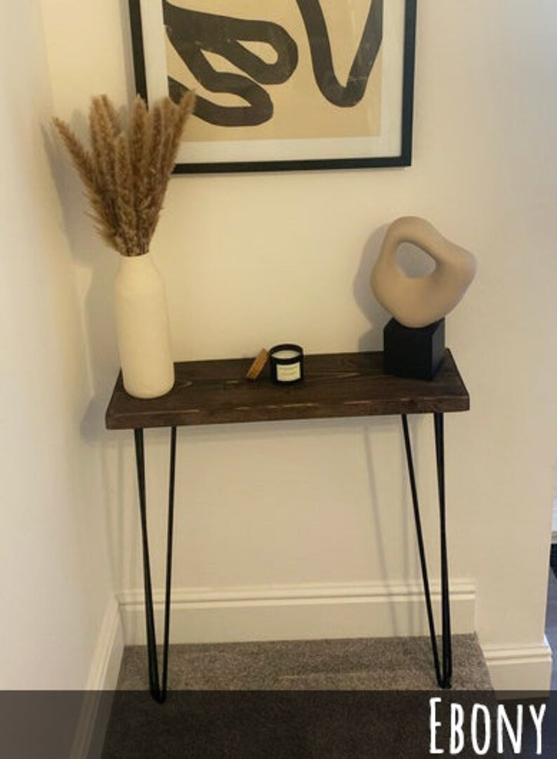 May include: A dark brown wooden console table with black hairpin legs. The table is decorated with a beige vase holding dried plants, a small candle, and a beige abstract sculpture on a black base. A framed abstract artwork hangs above.