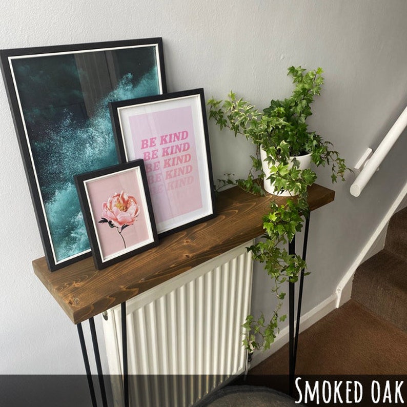 May include: A wooden console table with black hairpin legs, holding framed art and a potted plant. The art includes a floral print, an abstract ocean wave print, and a pink print with the words "BE KIND" repeated. The table is made of smoked oak.