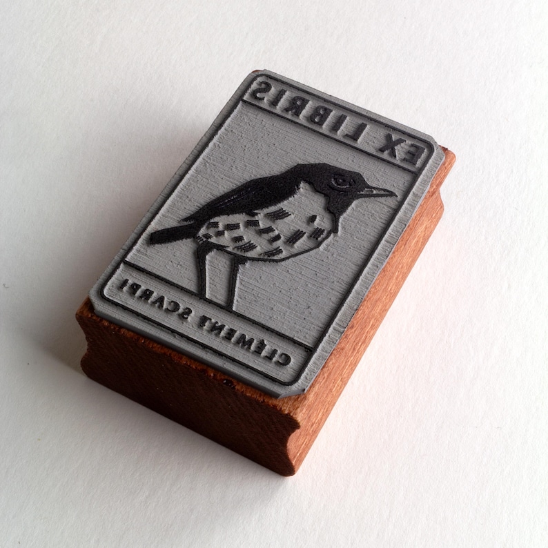A rubber stamp with an illustration of a bird and the text EX LIBRIS CLEMENT SCARPI rests beside its stamped impression on a book page, making a parfait cadeau fait main for collectors of bijoux uniques or vintage treasures.