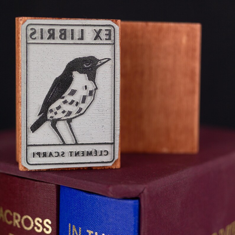 A rubber stamp with an illustration of a bird and the text EX LIBRIS CLEMENT SCARPI rests beside its stamped impression on a book page, making a parfait cadeau fait main for collectors of bijoux uniques or vintage treasures.