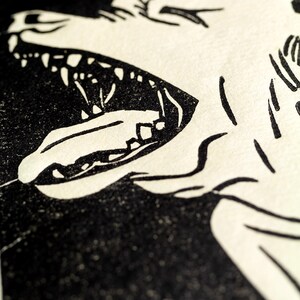 May include: Black and white illustration of a wolf's head with its mouth open, showing sharp teeth. The wolf's fur is detailed with lines and the background is black.