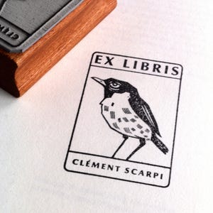 A rubber stamp with an illustration of a bird and the text EX LIBRIS CLEMENT SCARPI rests beside its stamped impression on a book page, making a parfait cadeau fait main for collectors of bijoux uniques or vintage treasures.