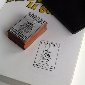 May include: A rectangular wooden rubber stamp with a metal plate featuring a beetle illustration and the text EX LIBRIS CLÉMENT SCARPI. The stamp is shown next to a black ink pad and a clear black ink impression on a white paper surface.