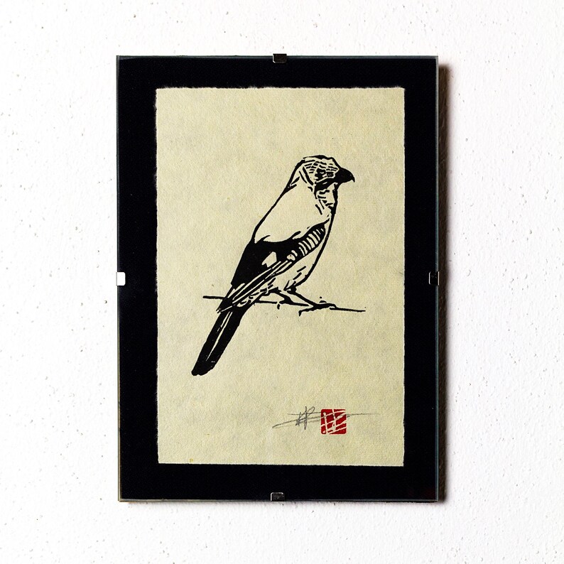May include: Framed print with a black border, showcasing a monochrome illustration of a bird. The bird is perched on a branch, rendered in a minimalist style with black lines. A red square stamp is in the lower right corner. The artwork is set against a cream-colored backdrop.