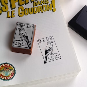 May include: A rectangular stamp featuring a bird illustration and the text "EX LIBRIS" and "CLEMENT SCARPI". The stamp is on a white surface with a printed image of the stamp. A black ink pad is in the background.