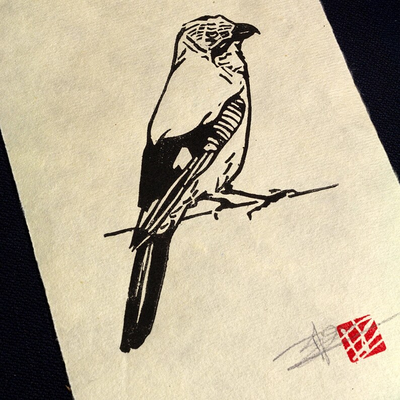 May include: A monochrome linocut print featuring a bird perched on a branch. The bird is rendered in a minimalist style with strong lines and a textured effect. A red square stamp is in the lower right corner. The print is on cream-coloured paper.