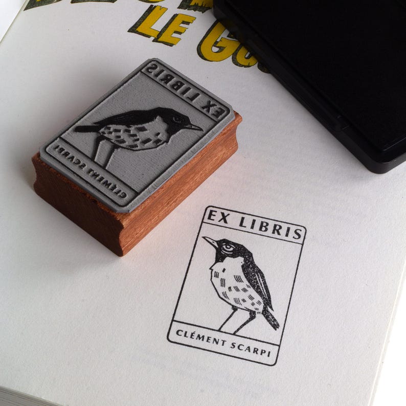 A rubber stamp with an illustration of a bird and the text EX LIBRIS CLEMENT SCARPI rests beside its stamped impression on a book page, making a parfait cadeau fait main for collectors of bijoux uniques or vintage treasures.
