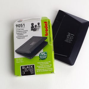 A black Trodat 9051 stamp pad sits next to its green and white box, which highlights features like 95% recycled plastic—perfect for packaging vêtements vintage, bijoux uniques, or any cadeau fait main. The product info appears in English, German, and Russian.