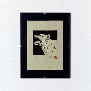May include: Black and white print of a dog with its mouth open, framed in a black frame. The print is on a cream-colored paper with a red stamp in the bottom right corner.