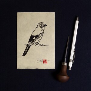 May include: A linocut print of a bird in black and white, on textured, off-white paper. A white mechanical pencil and a wood carving tool are next to the print. A red square stamp is in the lower right corner. The paper has a rough edge.