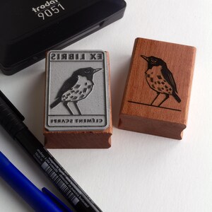 A rubber stamp with an illustration of a bird and the text EX LIBRIS CLEMENT SCARPI rests beside its stamped impression on a book page, making a parfait cadeau fait main for collectors of bijoux uniques or vintage treasures.