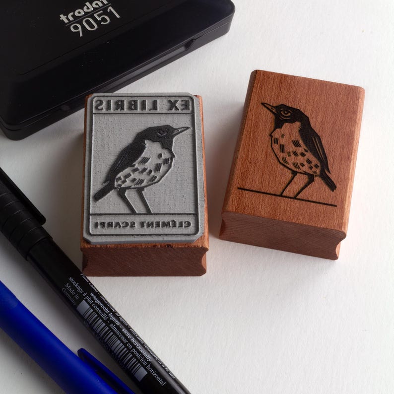A rubber stamp with an illustration of a bird and the text EX LIBRIS CLEMENT SCARPI rests beside its stamped impression on a book page, making a parfait cadeau fait main for collectors of bijoux uniques or vintage treasures.