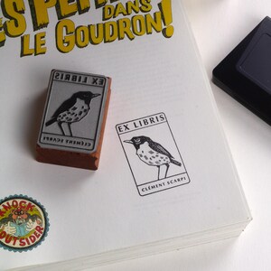 A rubber stamp with an illustration of a bird and the text EX LIBRIS CLEMENT SCARPI rests beside its stamped impression on a book page, making a parfait cadeau fait main for collectors of bijoux uniques or vintage treasures.