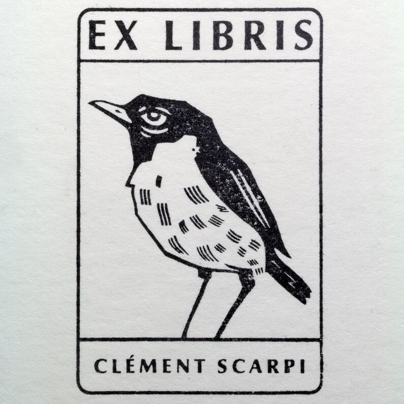 A rubber stamp with an illustration of a bird and the text EX LIBRIS CLEMENT SCARPI rests beside its stamped impression on a book page, making a parfait cadeau fait main for collectors of bijoux uniques or vintage treasures.