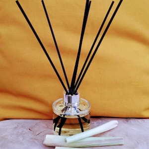 May include: A clear glass bottle with a silver cap and black diffuser sticks. The bottle is filled with a light yellow liquid. There are four pieces of dried lemongrass on a wooden surface.