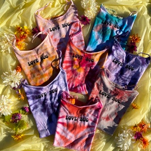 May include: Eight tie-dye tank tops with adjustable straps. Each tank top features the text "LOVE BUG" in black lettering.