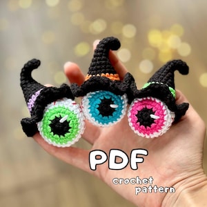Puede incluir: Tres adornos de ojo tejidos a crochet con sombreros de bruja negros. Los ojos son verdes, azules y rosas. El texto "PDF crochet pattern" está debajo de los adornos.