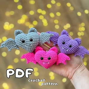 May include: PDF crochet pattern for making three heart-shaped bats. The bats are gray, purple, and pink. The bats have black eyes and white fangs.