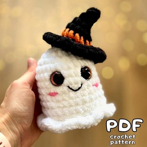Könnte beinhalten: Ein weißer gehäkelter Geist, der einen schwarzen Hexenhut mit orangefarbenem Besatz trägt. Der Geist hat große braune Augen und ein rosafarbenes Lächeln. Der Text "PDF crochet pattern" ist in der rechten unteren Ecke des Bildes zu sehen.
