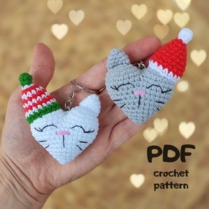 May include: Two crocheted heart-shaped cat keyrings with a festive holiday theme. One cat is white with a red and green striped hat, the other is grey with a red Santa hat. The text "PDF crochet pattern" is visible below the keyrings.