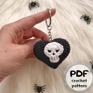 Puede incluir: Un llavero negro en forma de corazón tejido a ganchillo con un diseño de calavera blanca. El llavero está unido a un anillo y una cadena de metal plateado. La imagen también incluye un gráfico de patrón de ganchillo PDF.