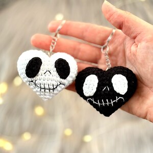 May include: Two crocheted heart-shaped keychains with skull faces. One keychain is white with black eyes and a black stitched mouth. The other keychain is black with white eyes and a white stitched mouth.