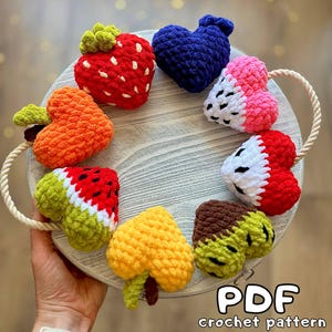 May include: A collection of colorful, crocheted fruit and heart shapes arranged on a round wooden tray with a rope handle. The fruits include a red strawberry, orange carrot, watermelon, and a yellow leaf. The text "PDF crochet pattern" is visible.