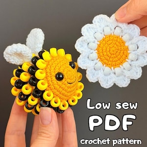Puede incluir: Una abeja de ganchillo con cuentas de plástico amarillas y negras y alas blancas de ganchillo, junto a una margarita de ganchillo con centro amarillo y pétalos blancos con cuentas blancas. Texto: Low sew PDF crochet pattern.