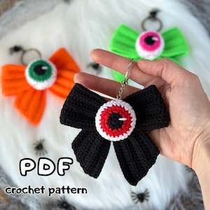 May include: Crocheted Halloween-themed keychains in orange, green, and black bow shapes, each with a central eyeball design. The keychains are on a white fluffy surface. The text "PDF crochet pattern" is visible.