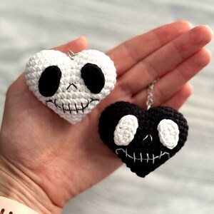 May include: Two crocheted heart-shaped keychains with a skull design. One keychain is white with black eyes and a black stitched mouth. The other keychain is black with white eyes and a white stitched mouth.