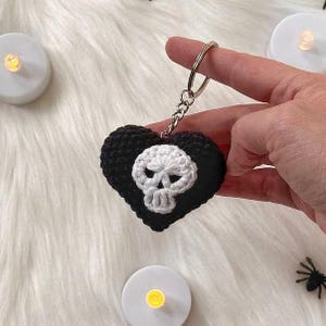 Puede incluir: Un llavero negro en forma de coraz&oacute;n tejido a ganchillo con un dise&ntilde;o de calavera blanca. El llavero se sujeta con un anillo plateado. La calavera est&aacute; centrada en el coraz&oacute;n. El fondo es una superficie blanca y esponjosa.