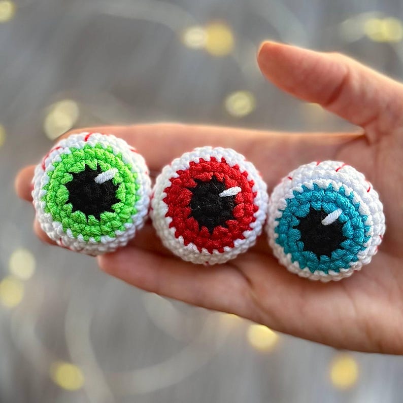 May include: Three crocheted eyeballs, each with a different colored iris: green, red, and blue. The eyeballs are white with black pupils and are made with a textured yarn.