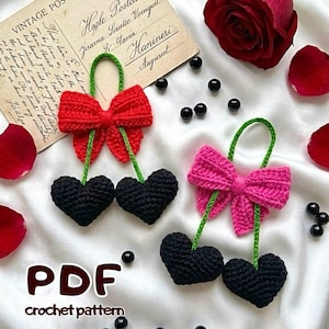 May include: Crocheted cherry ornaments with black heart-shaped cherries and red and pink bows. The cherries have green stems and loops. The image includes a vintage postcard, rose petals, and the text "PDF crochet pattern."