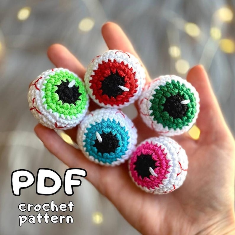May include: Five crocheted eyeball ornaments in different colors: red, green, blue, pink, and white. The eyeballs have black pupils and white highlights. The text "PDF crochet pattern" is written below the eyeballs.