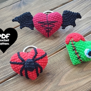 Puede incluir: Tres llaveros de corazón de ganchillo con diferentes diseños. Un corazón tiene alas de murciélago negras, otro tiene una araña negra y el tercero tiene un corazón verde cosido con un ojo saltones.