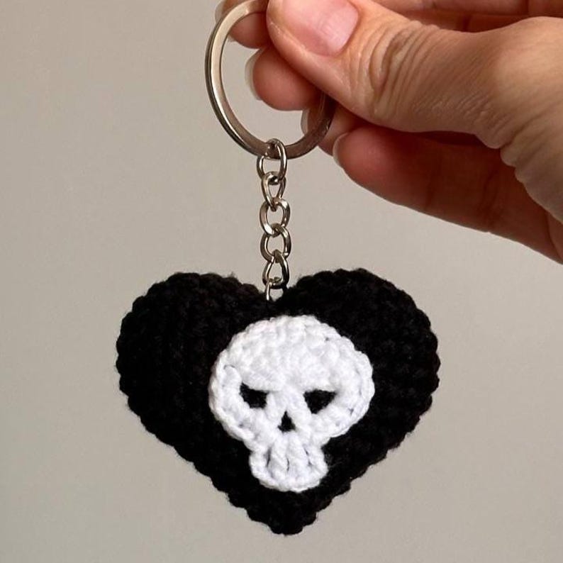 Puede incluir: Un llavero negro en forma de coraz&oacute;n tejido a ganchillo con un dise&ntilde;o de calavera blanca en el centro. El llavero tiene un anillo y una cadena de metal plateado. El coraz&oacute;n est&aacute; sostenido por una mano.