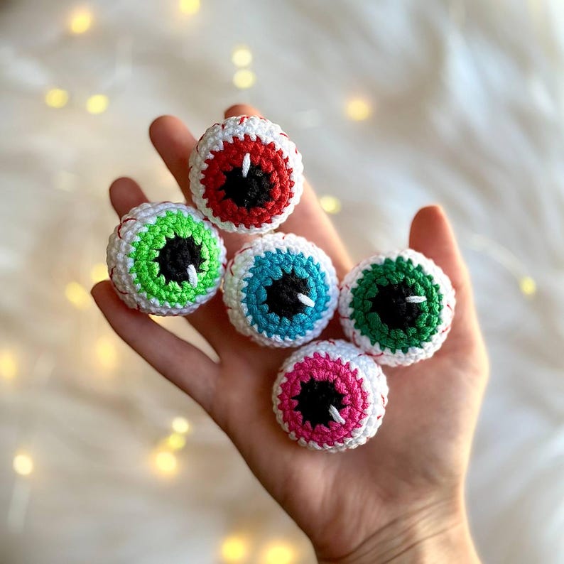May include: Five crocheted eyeball ornaments in different colors. The eyeballs are white with a black pupil and a colored iris. The colors are red, green, blue, pink, and green.