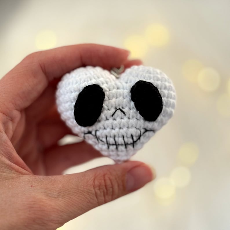 May include: White crocheted heart-shaped ornament with a black skull face. The skull has two black eyes and a stitched-on smile.