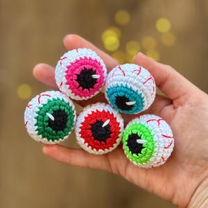 May include: Five crocheted eyeball ornaments in different colors. The eyeballs are white with red veins and have different colored irises: pink, blue, green, red, and green.