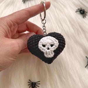 Puede incluir: Un llavero en forma de coraz&oacute;n negro y blanco tejido a ganchillo con un dise&ntilde;o de calavera. El llavero est&aacute; sujeto por una mano y presenta un anillo plateado. La calavera es blanca y el coraz&oacute;n es negro. El fondo es una superficie blanca y esponjosa.