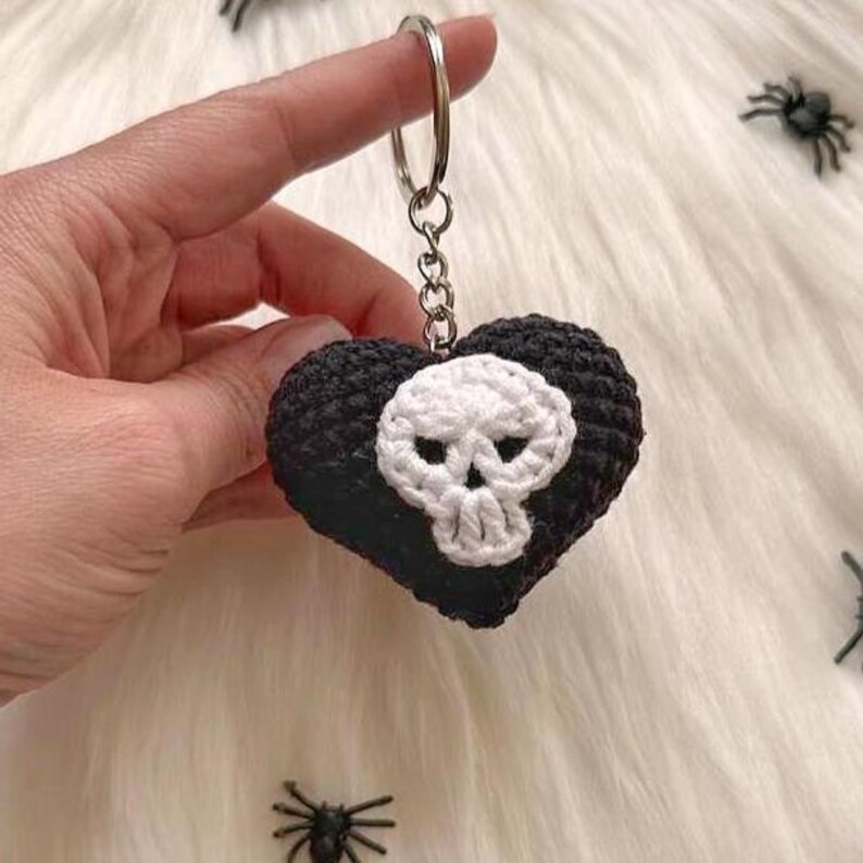 Puede incluir: Un llavero en forma de coraz&oacute;n negro y blanco tejido a ganchillo con un dise&ntilde;o de calavera. El llavero est&aacute; sujeto por una mano y presenta un anillo plateado. La calavera es blanca y el coraz&oacute;n es negro. El fondo es una superficie blanca y esponjosa.