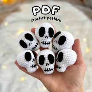 May include: A collection of six small, crocheted skulls in white with black eye sockets and stitched mouths. The image includes the text "PDF crochet pattern" in a playful font, suggesting a DIY craft project.