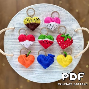 May include: A selection of crochet heart keyrings, each designed to resemble different fruits such as strawberries and watermelons. The keyrings are displayed on a white wooden tray. The text "PDF crochet pattern" is visible. Each keyring has a metal ring.